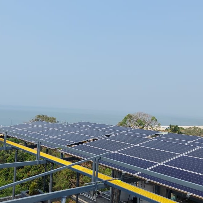 Rooftop solar panels under a clear blue sky, with a distant view of the ocean and greenery. Bright, sunny day, conveying sustainability and nature.