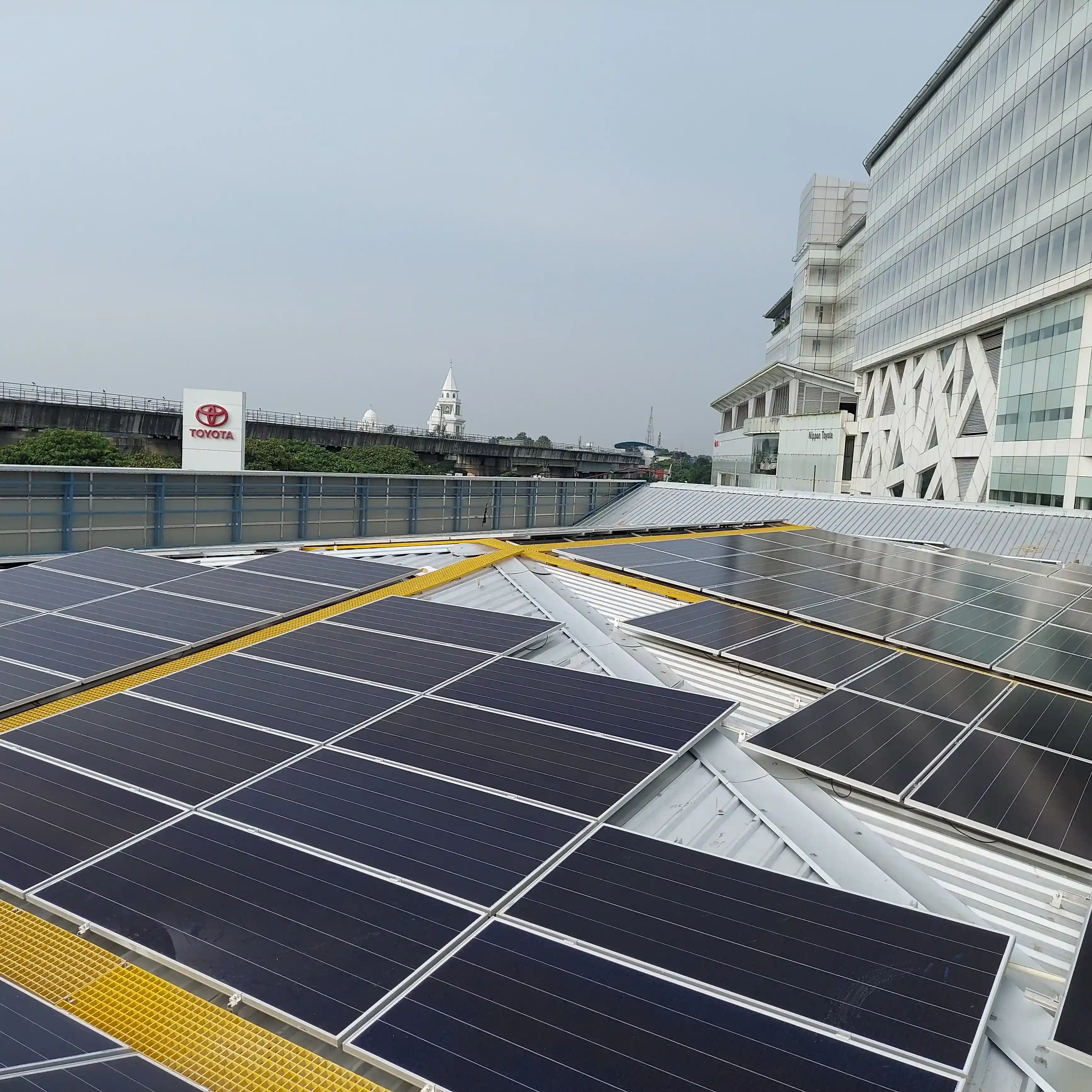 Rooftop solar panels under a clear sky, with a modern glass building and a distant tower. An overpass with green foliage is visible on the left.