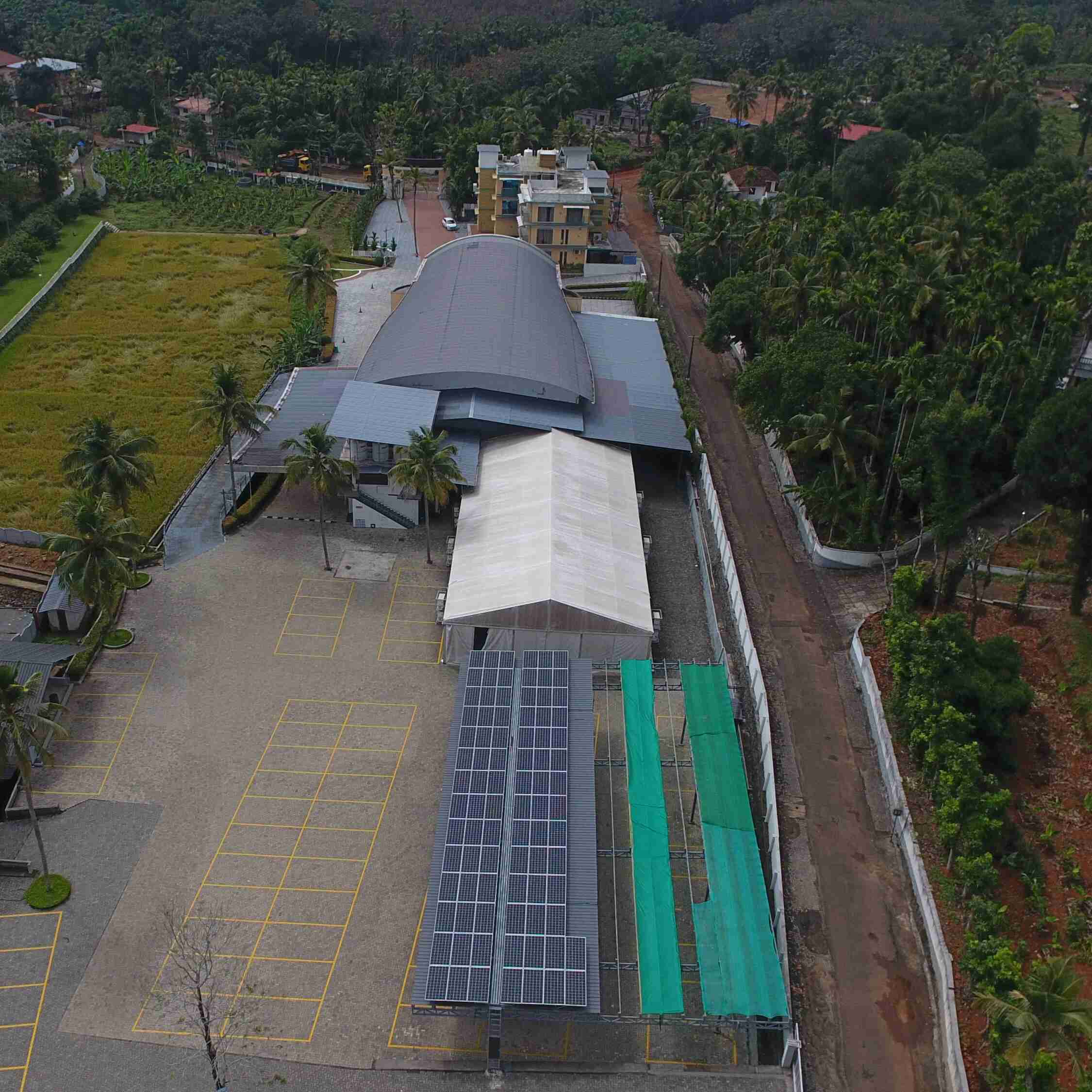Aerial view of an expansive building with a curved roof, surrounded by lush greenery and fields. Solar panels are visible on one side, conveying sustainability.