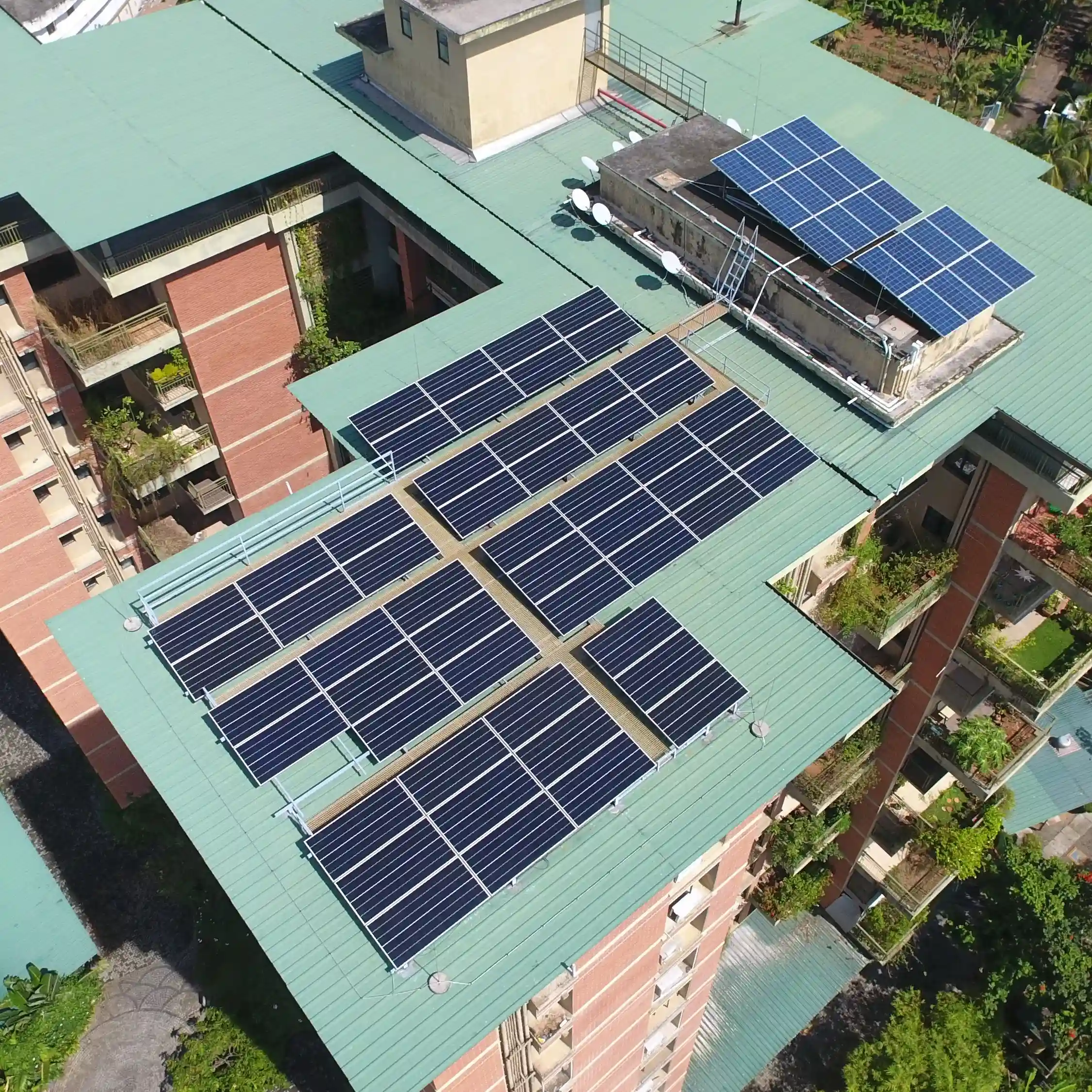 Aerial view of a large building with its rooftop covered in multiple solar panels, surrounded by lush greenery. The green metal roof contrasts with brick walls, emphasizing sustainable architecture.