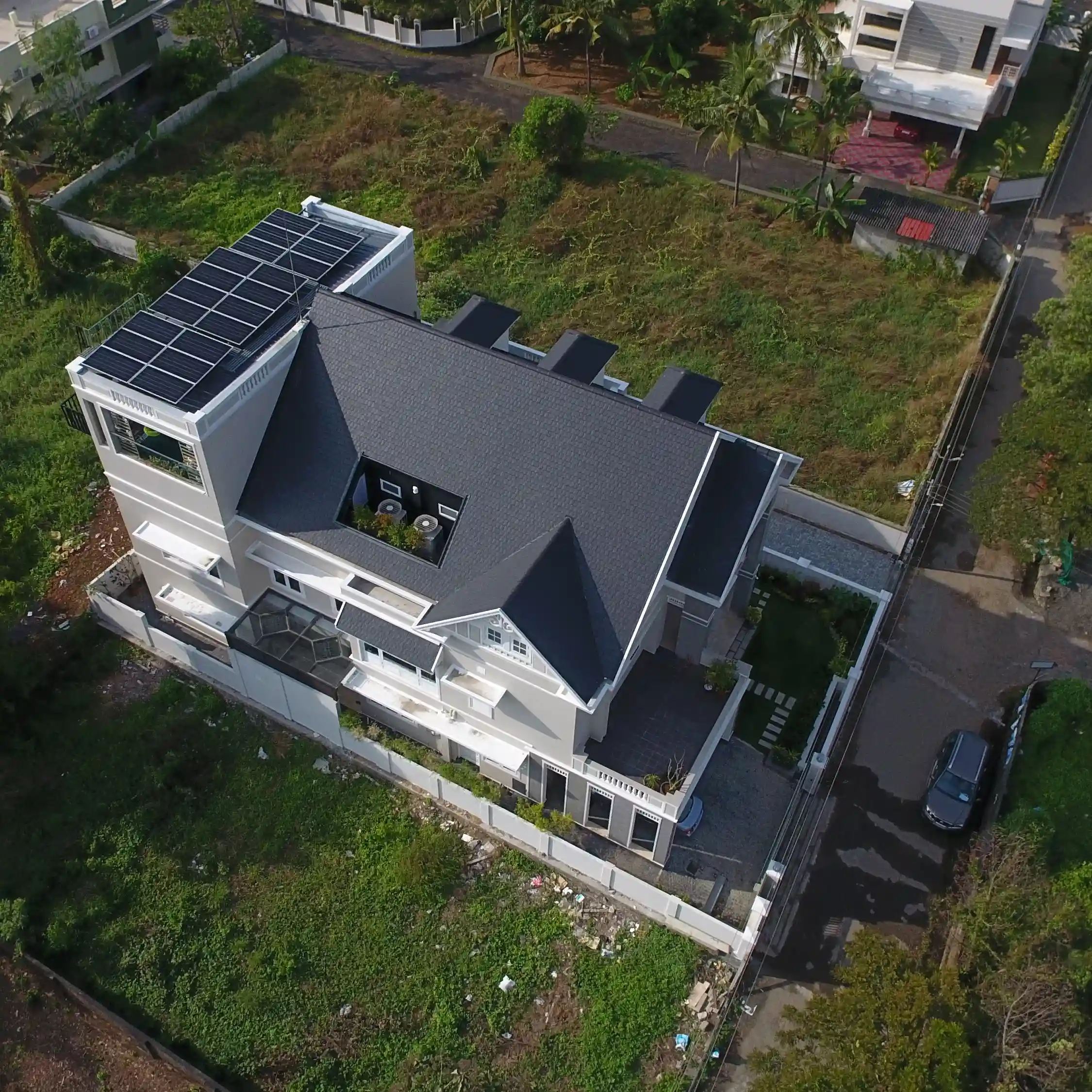 Aerial view of a modern house with a dark roof, solar panels, and a courtyard. Surrounded by greenery and other houses, with a car on a nearby road.