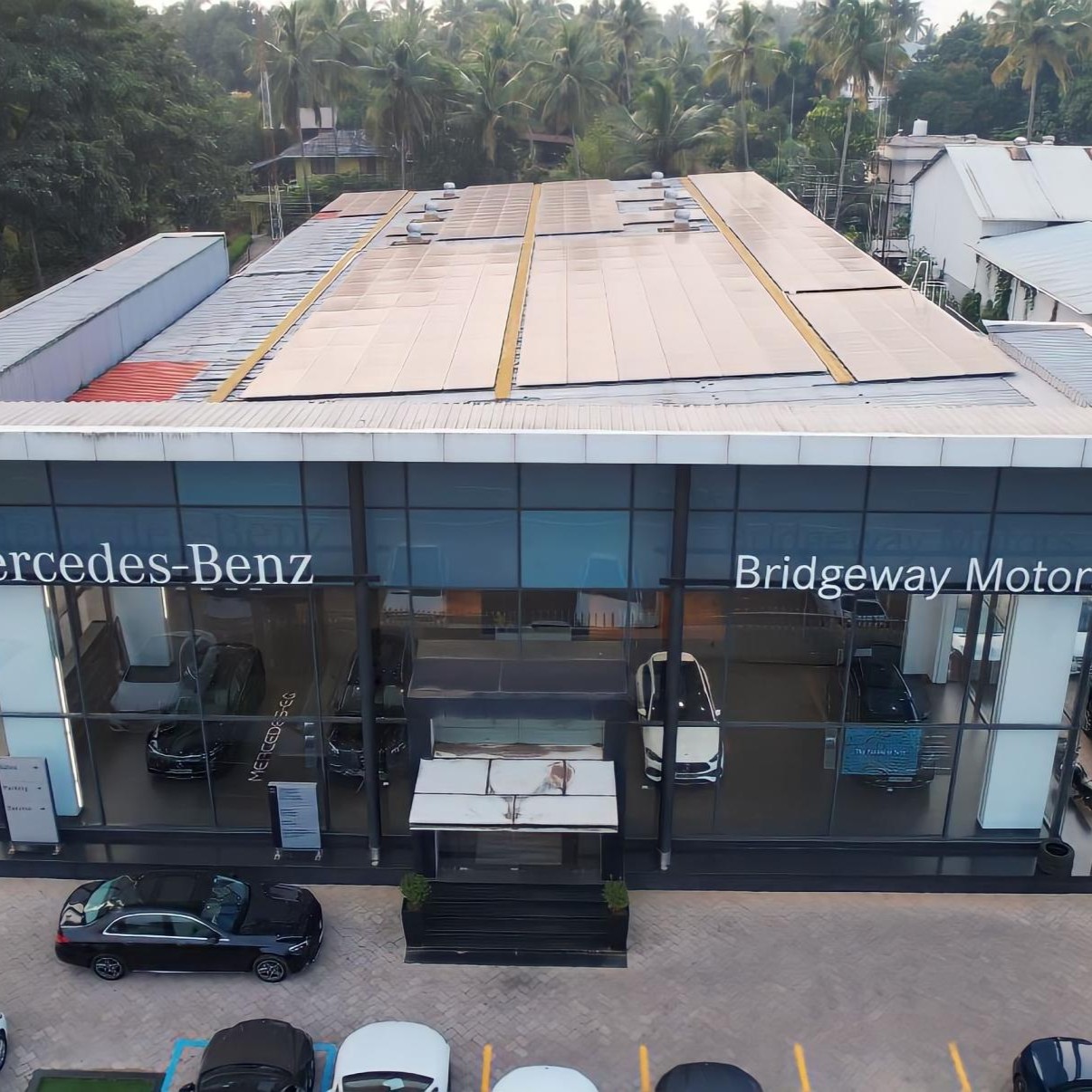 Aerial view of a Mercedes-Benz showroom surrounded by lush greenery. The glass facade displays cars inside. The atmosphere is serene and modern.